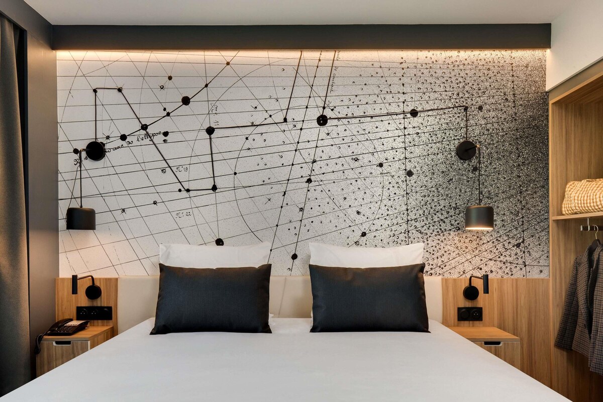 A modern bedroom with a geometric wall design, two bedside tables with lamps, a bed with two pillows, and a wooden wardrobe.