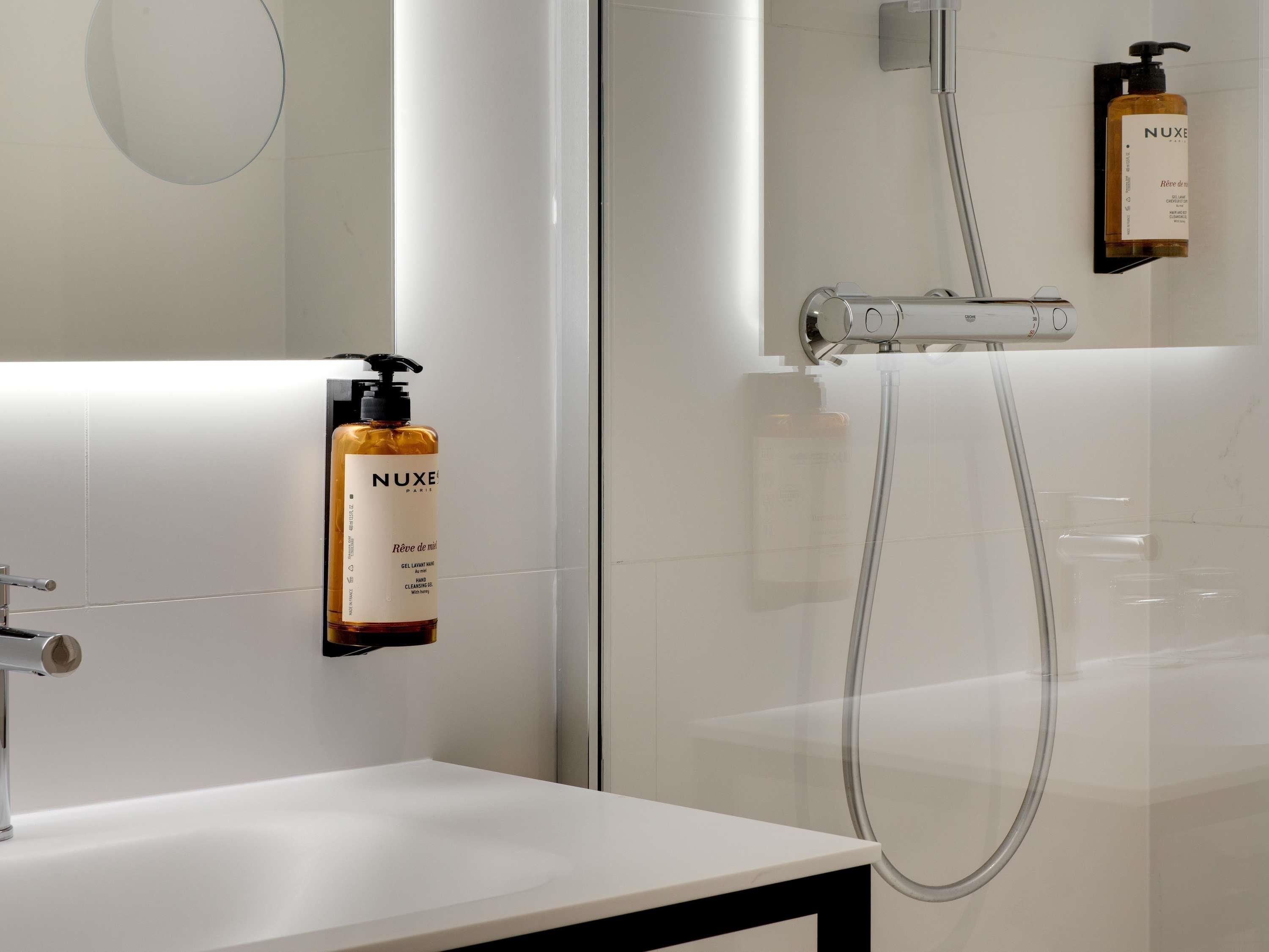 Une salle de bain moderne avec un lavabo blanc, une douchette à main et deux flacons de shampoing Nuxe Paris fixés au mur.