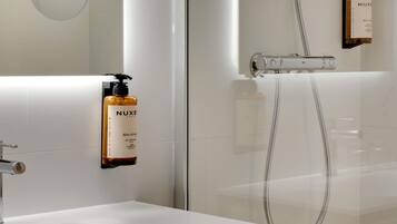 Une salle de bain moderne avec un lavabo blanc, une douchette à main et deux flacons de shampoing Nuxe Paris fixés au mur.