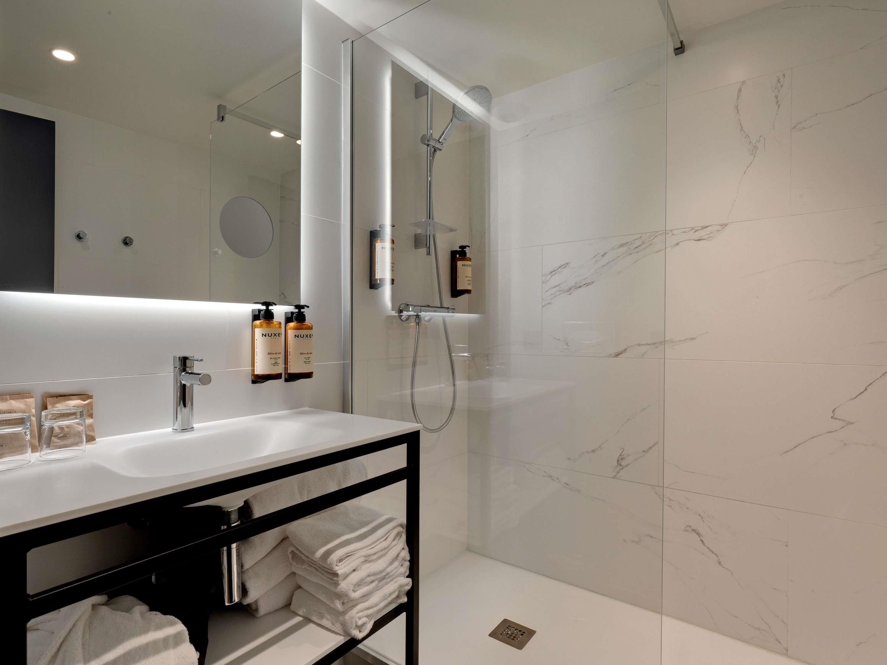A modern bathroom with a white marble wall, a large mirror, a sink with a black shelf, and a shower area.