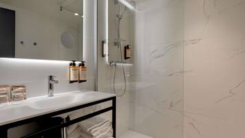 A modern bathroom with a white marble wall, a large mirror, a sink with a black shelf, and a shower area.