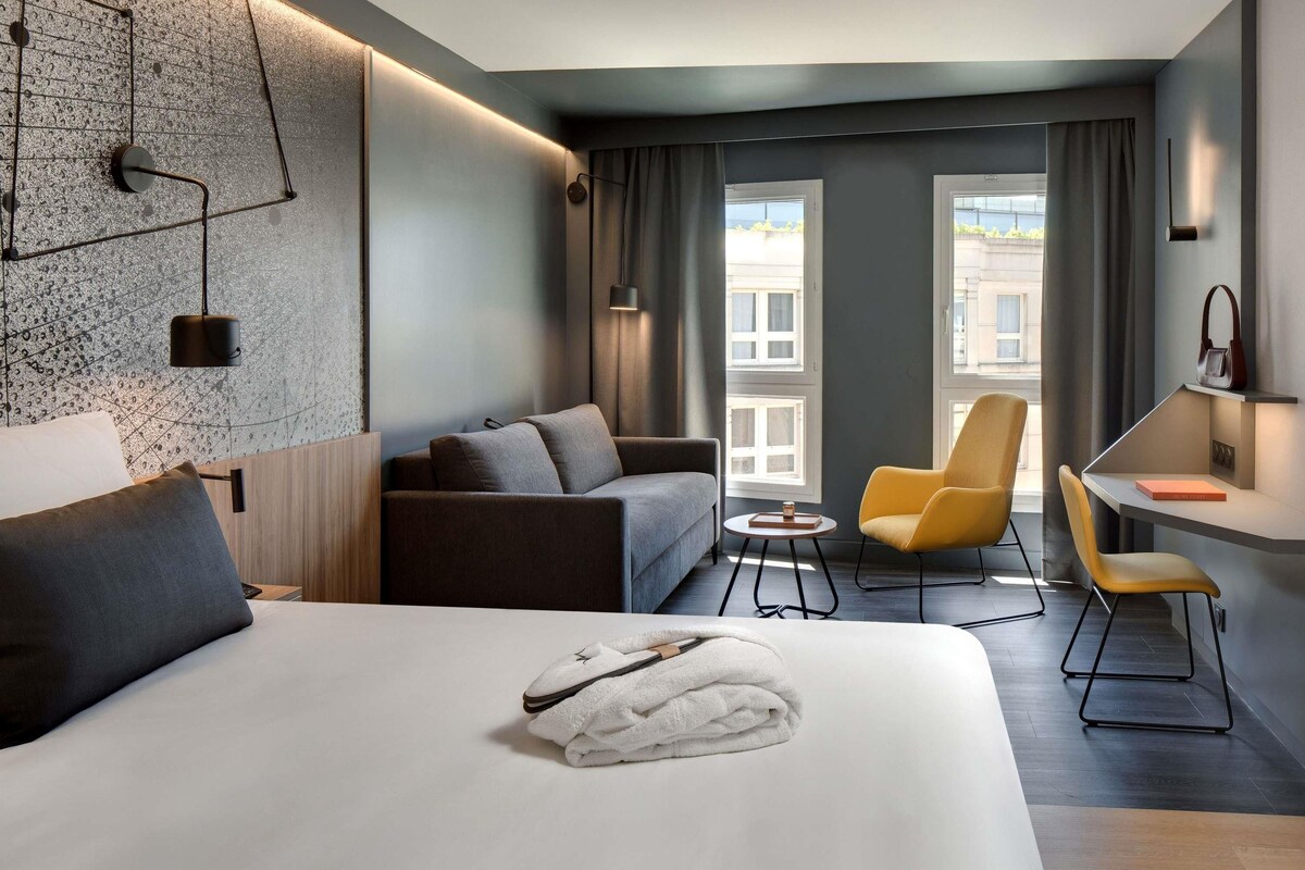 A modern hotel room with a large bed, a sofa, a chair, a small table, and a desk.