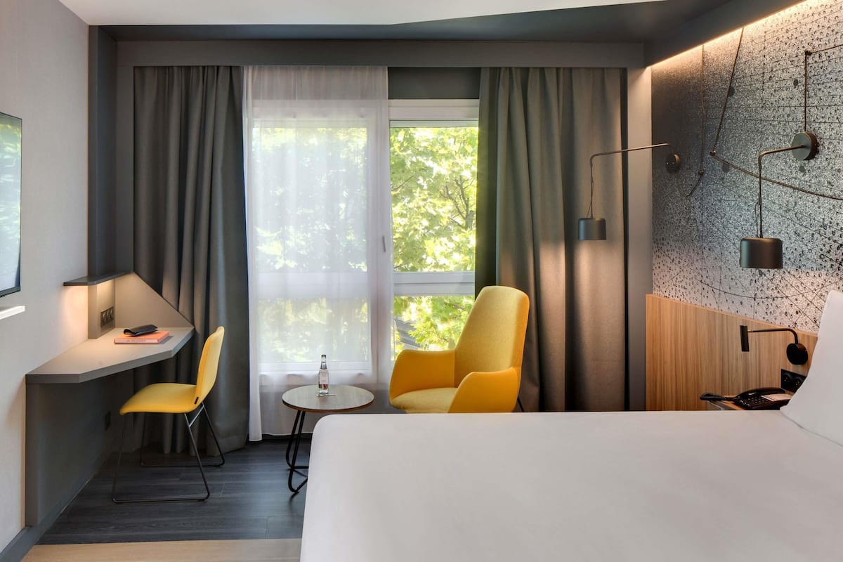 A hotel room with a bed, a desk, two chairs, a small table, a lamp, and a view of trees through the window.