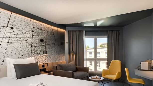A modern hotel room with a large bed, a sofa, a desk, and a chair. There is a window with curtains, a wall with a geometric pattern, and a view of buildings outside. - Mercure Montpellier Centre Antigone (Montpellier)