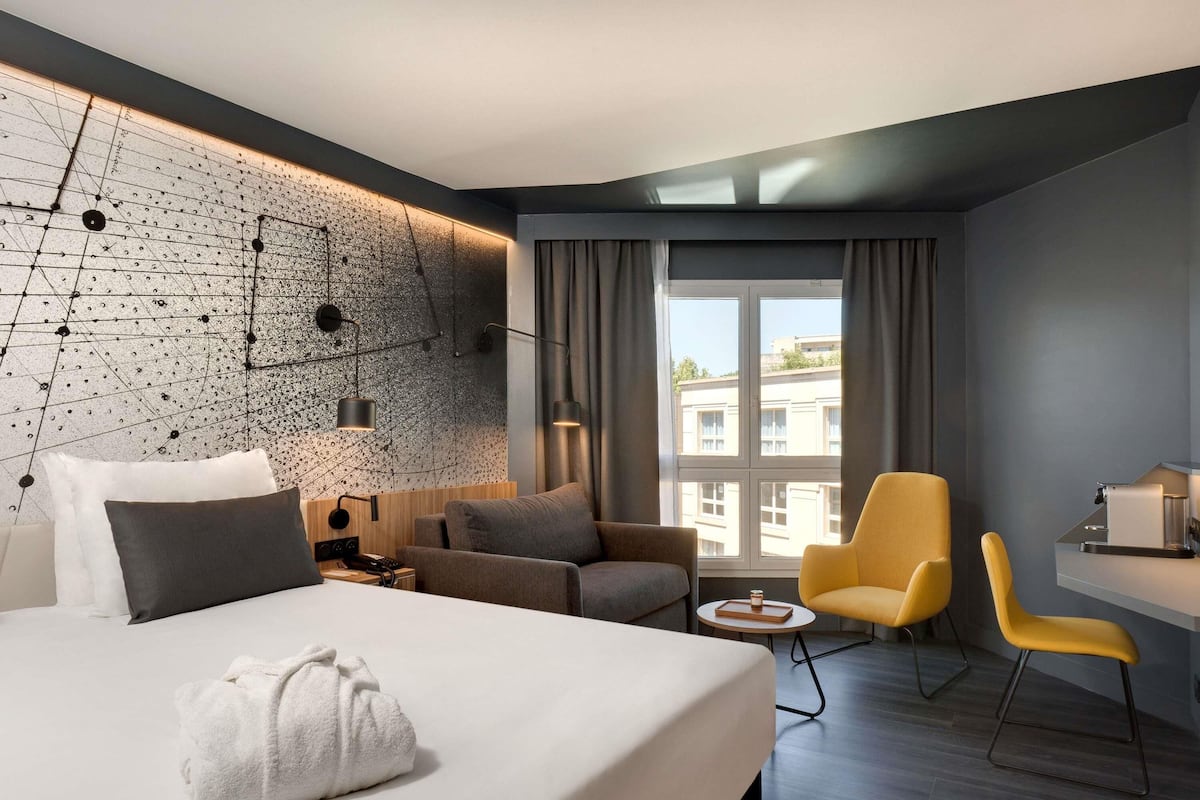 A modern hotel room with a large bed, a sofa, a desk, and a chair. There is a window with curtains, a wall with a geometric pattern, and a view of buildings outside.