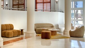 Un hall d’entrée spacieux avec un plafond haut, un espace de détente central agrémenté de canapés et de poufs, un sol en marbre et de grandes fenêtres équipées de stores.
