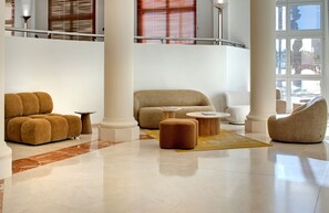 A spacious lobby with a high ceiling, a central seating area with sofas and ottomans, a marble floor, and large windows with blinds. - Mercure Montpellier Centre Antigone (Montpellier)