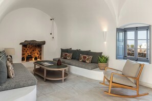A cozy living room with a stone fireplace, a grey sofa with patterned cushions, a wooden rocking chair, and a round coffee table.