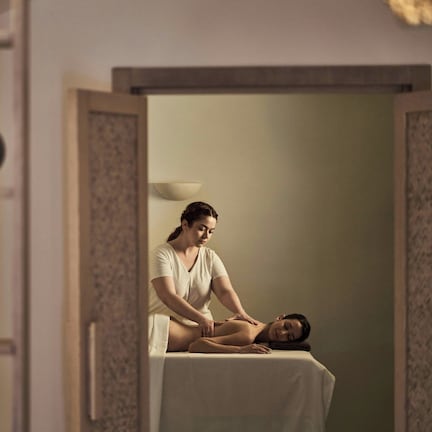 A massage therapy room with a client lying on a treatment table, a therapist attending to the client, and a decorative vase on a shelf.