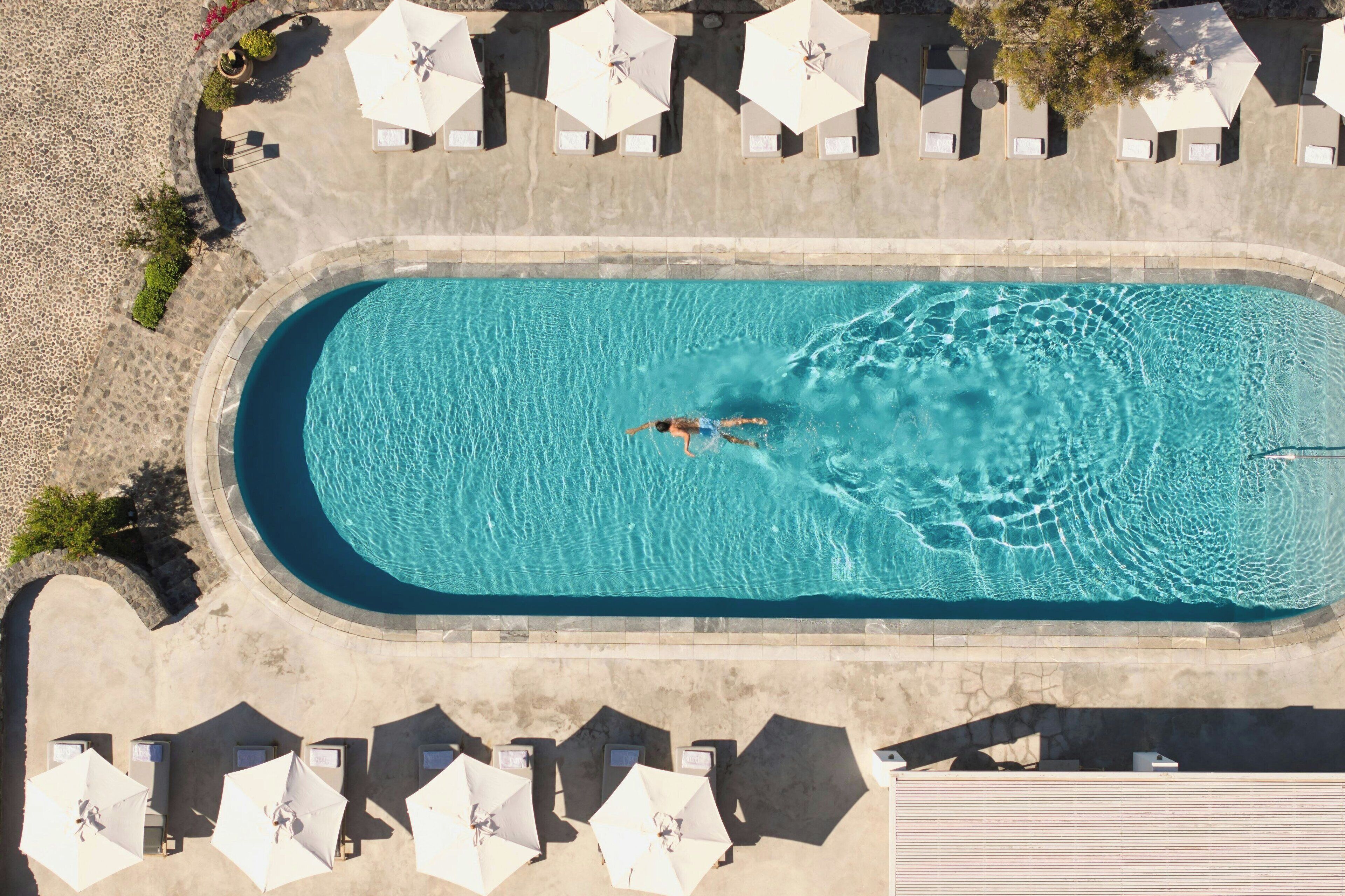 Luftaufnahme eines Schwimmbeckens mit einer darin schwimmenden Person, umgeben von Sonnenschirmen und Liegestühlen.