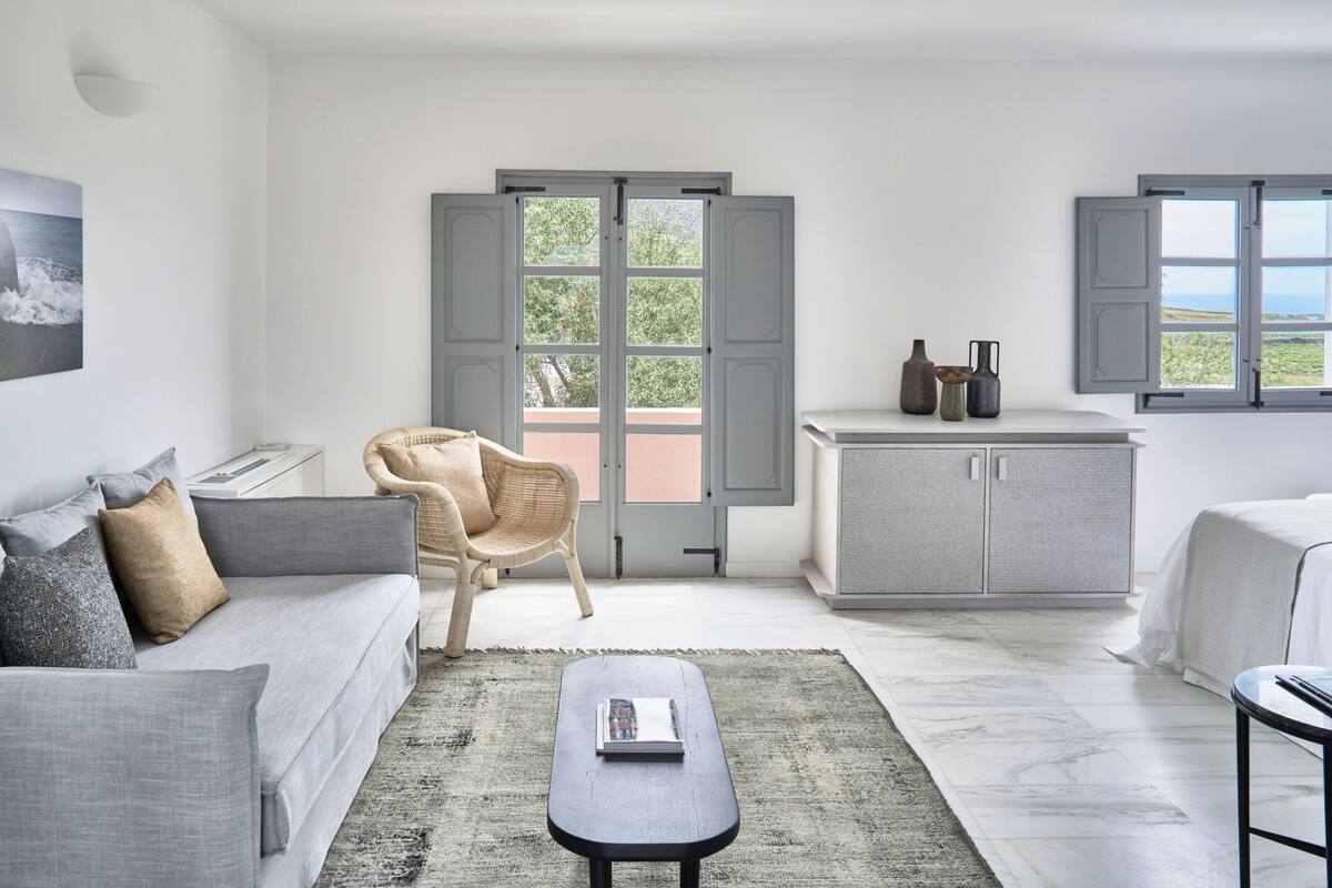 A modern, well-lit room with a grey sofa, a wicker chair, a small table with a book, a cabinet, and a bed.