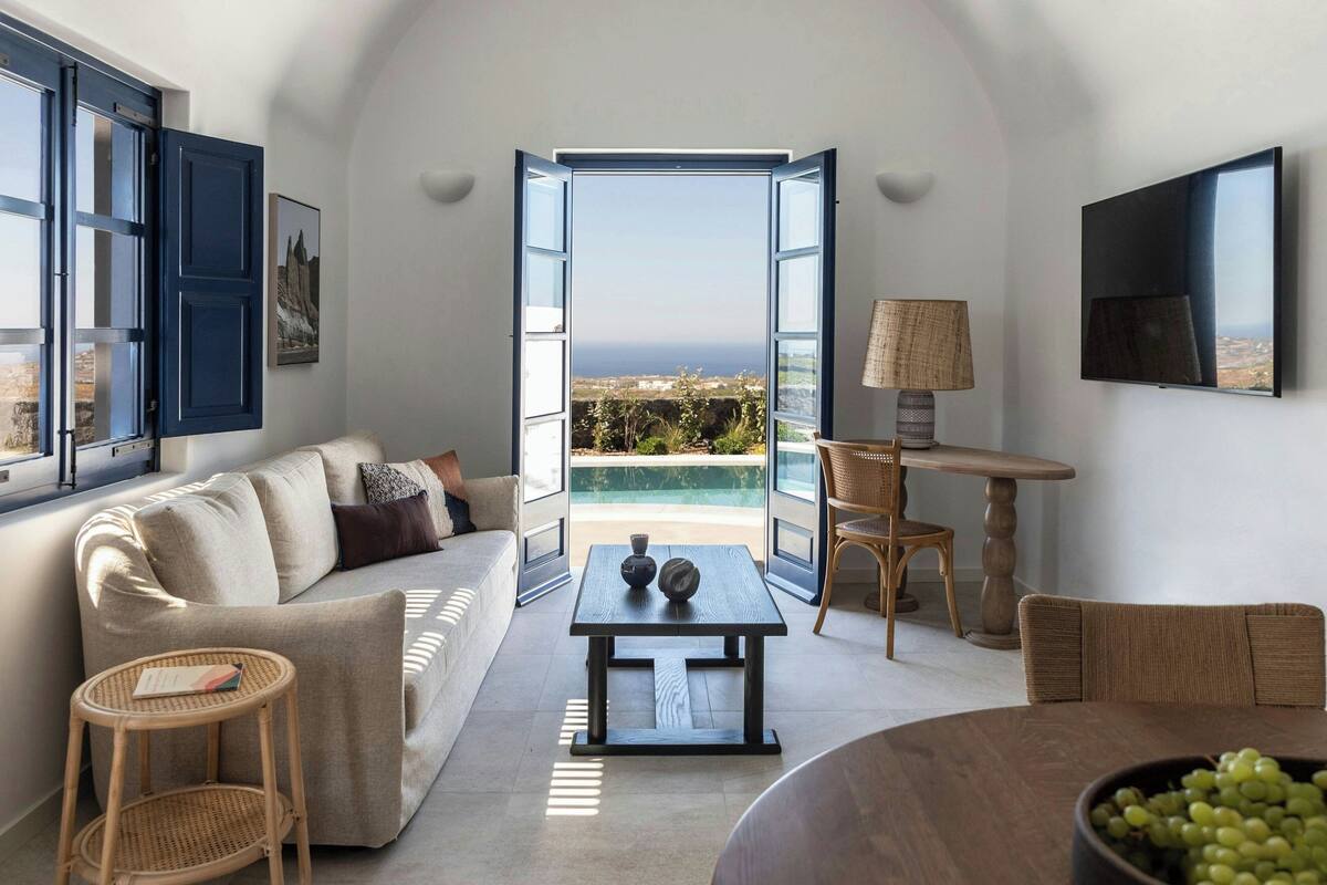 A modern living room with a sofa, a coffee table, a dining table with chairs, a TV mounted on the wall, and a large window with an open door leading to a balcony with a view of the sea.