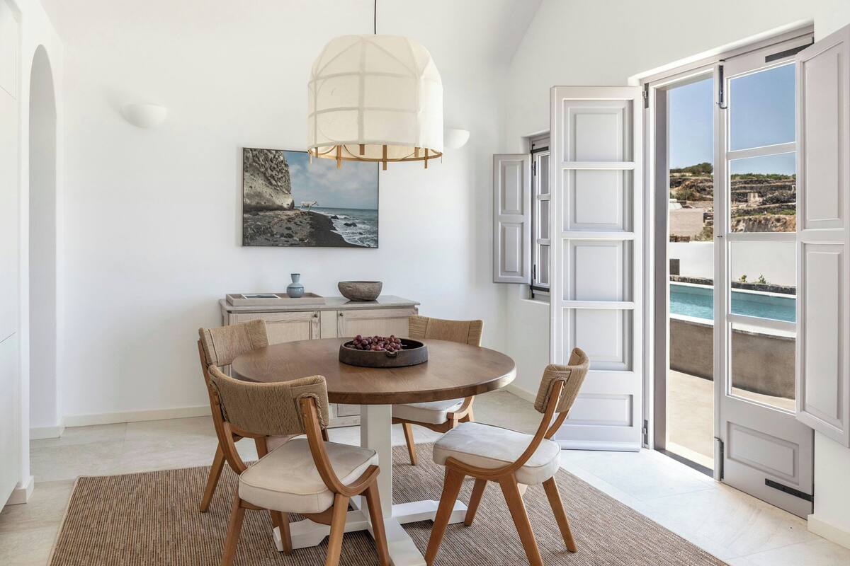 A dining area with a round table, four chairs, a hanging light fixture, a painting of a coastal scene, and a view of the sea through open doors.