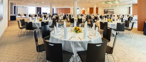 Een ruime balzaal met ronde tafels gedekt voor een diner, met witte tafelkleden en zwarte stoelen.