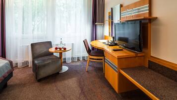 A hotel room with a bed, a desk with a TV, a chair, a small table with a bottle and apples, and a window with curtains.