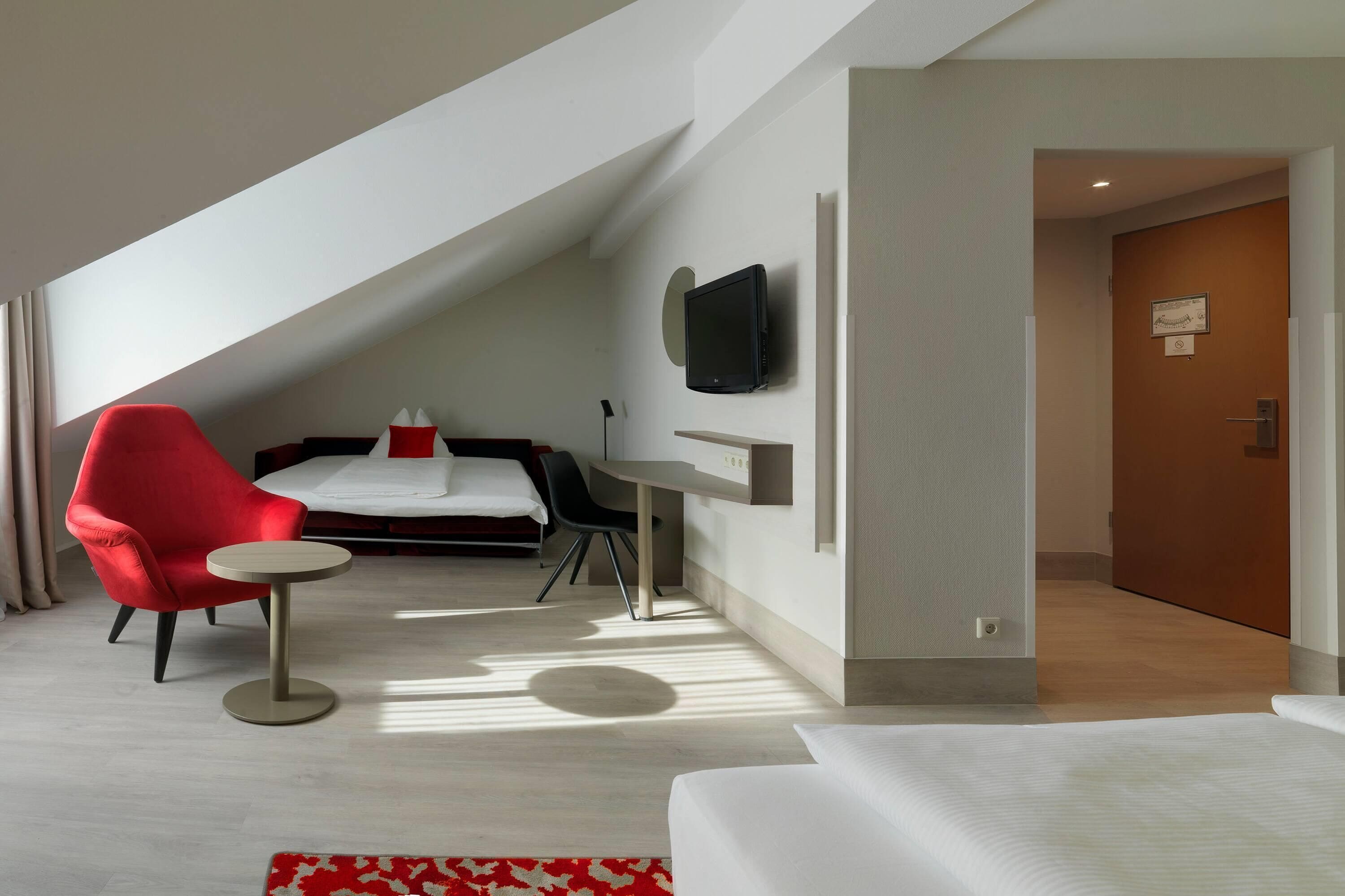 Una camera d'albergo moderna con soffitto spiovente, un letto con biancheria bianca, una poltrona rossa, un piccolo tavolino rotondo, un televisore a schermo piatto montato a parete e una porta in legno.