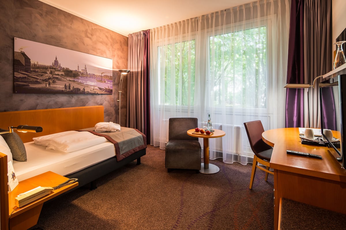 A hotel room with a bed, a desk, a chair, a small table, and a painting on the wall.