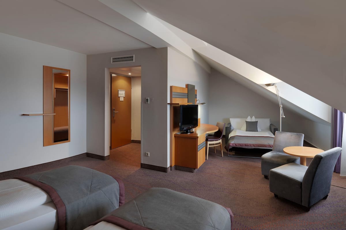 A hotel room with a bed, two armchairs, a small table, a TV, and a desk with a chair.