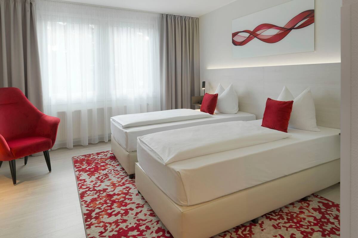 A hotel room with two beds, a red chair, a patterned rug, and a modern abstract painting on the wall.