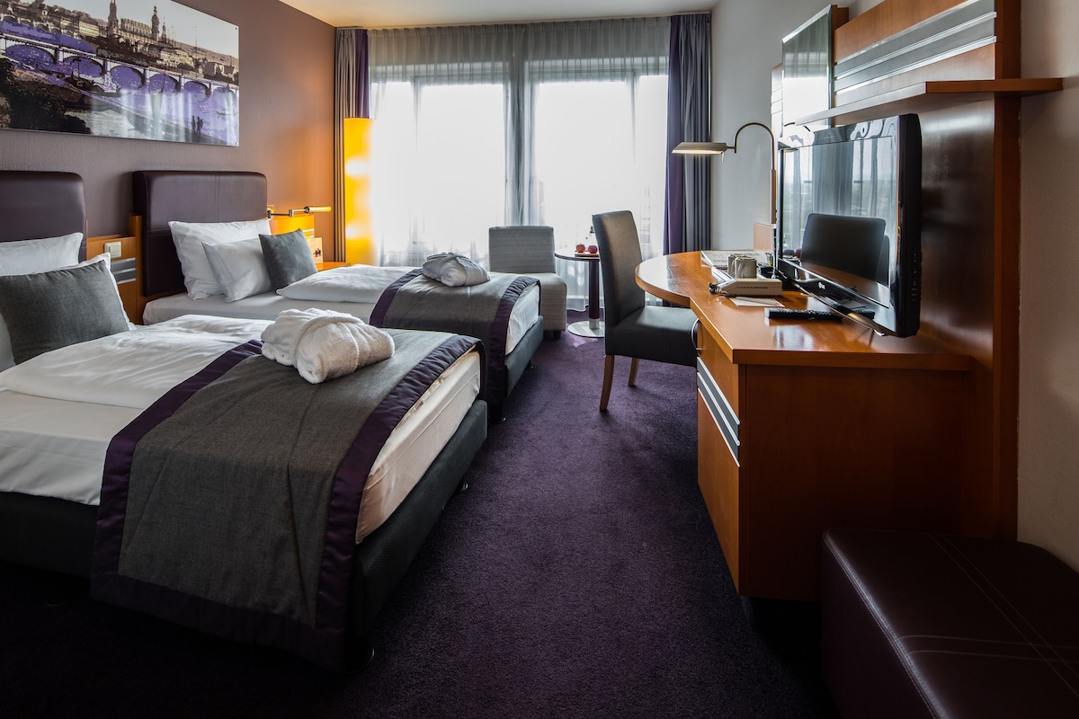 A hotel room with two beds, a desk with a television, a chair, and a view of a cityscape through large windows.