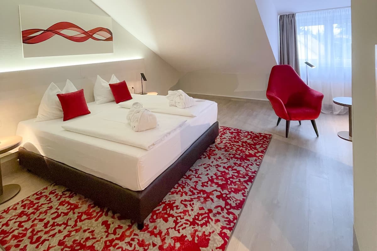 A modern hotel room with a large bed, a red chair, and a view of the city.