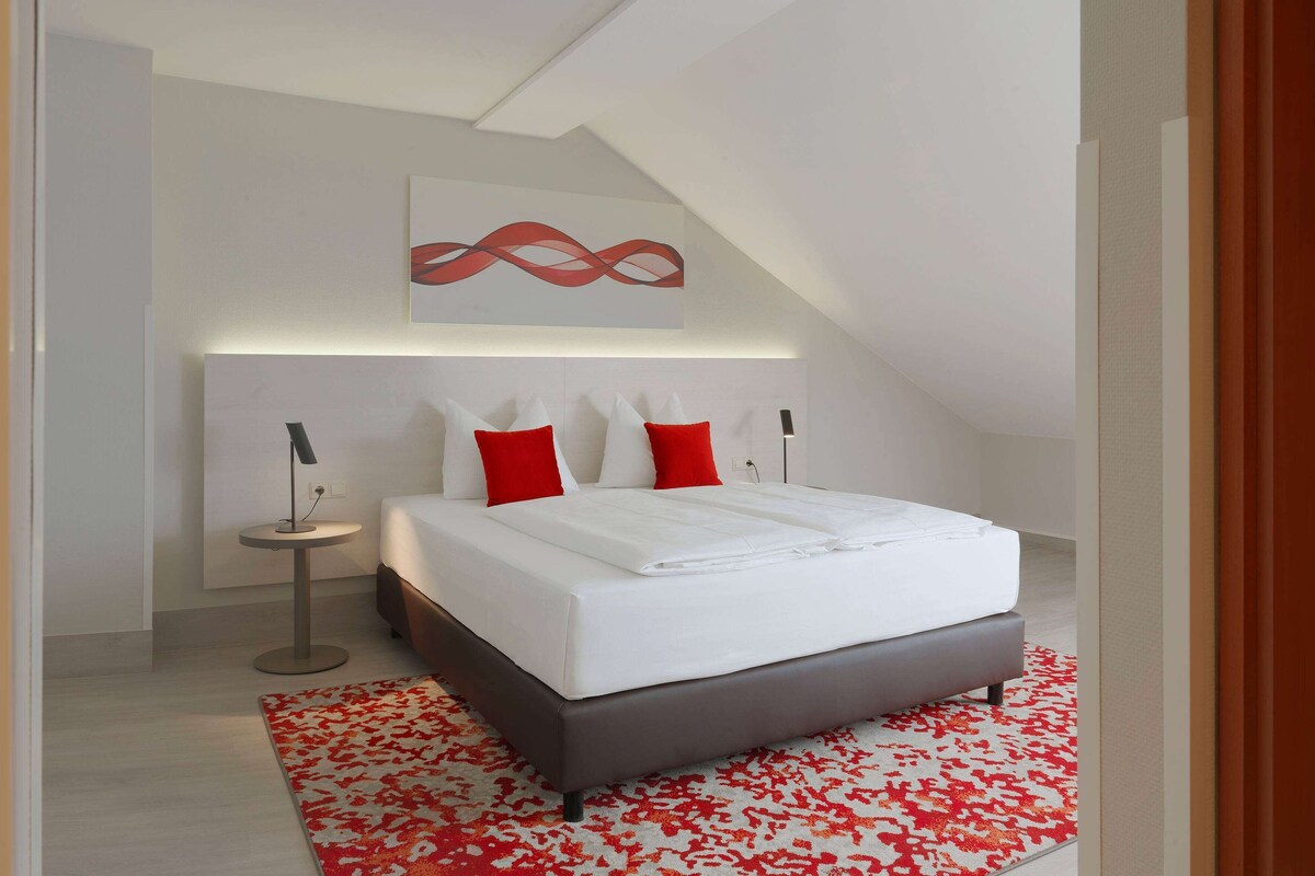 A modern bedroom with a bed, red and white pillows, a small bedside table, and a red and white patterned rug.