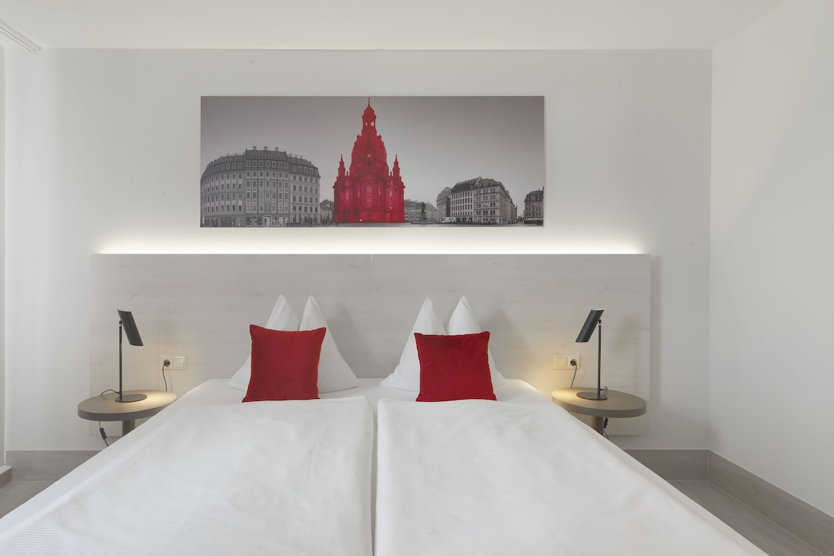 A hotel room with a large bed, two bedside tables with lamps, and a cityscape painting above the bed.