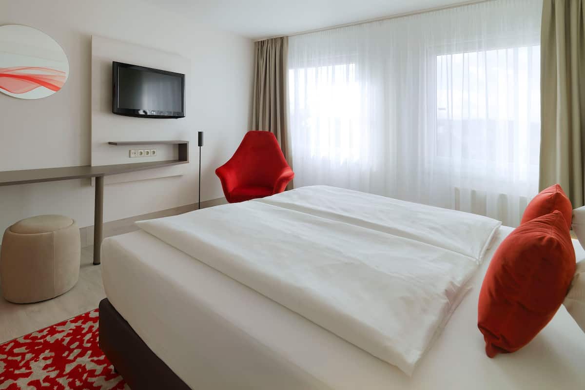 A hotel room with a bed, a red chair, a television mounted on the wall, and a window with curtains.