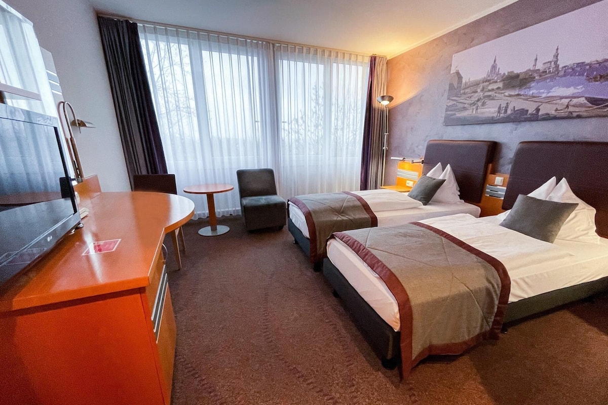 A hotel room with two beds, a desk, a chair, a small table, and a painting on the wall.
