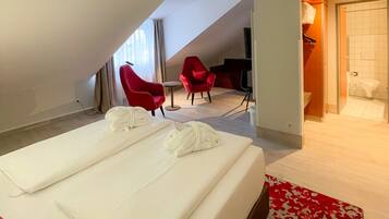 Une chambre d’hôtel avec un lit, deux chaises rouges, une petite table et une salle de bain visible à travers une porte ouverte.