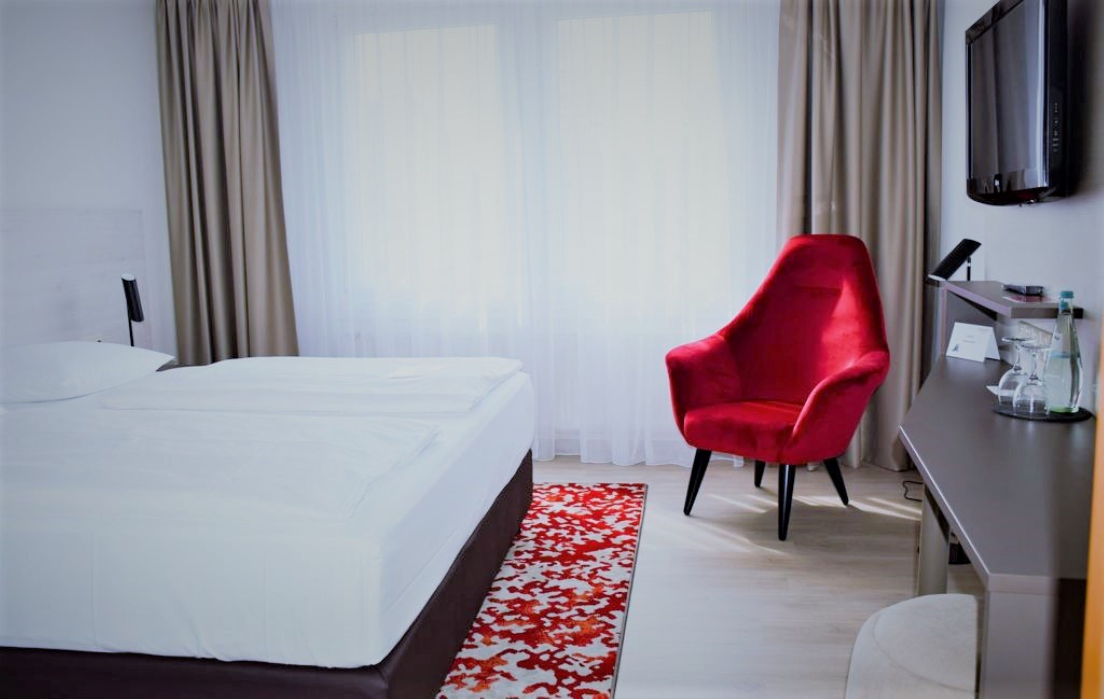 Una habitación de hotel con cama, una silla roja, un escritorio con televisor y una ventana con cortinas.