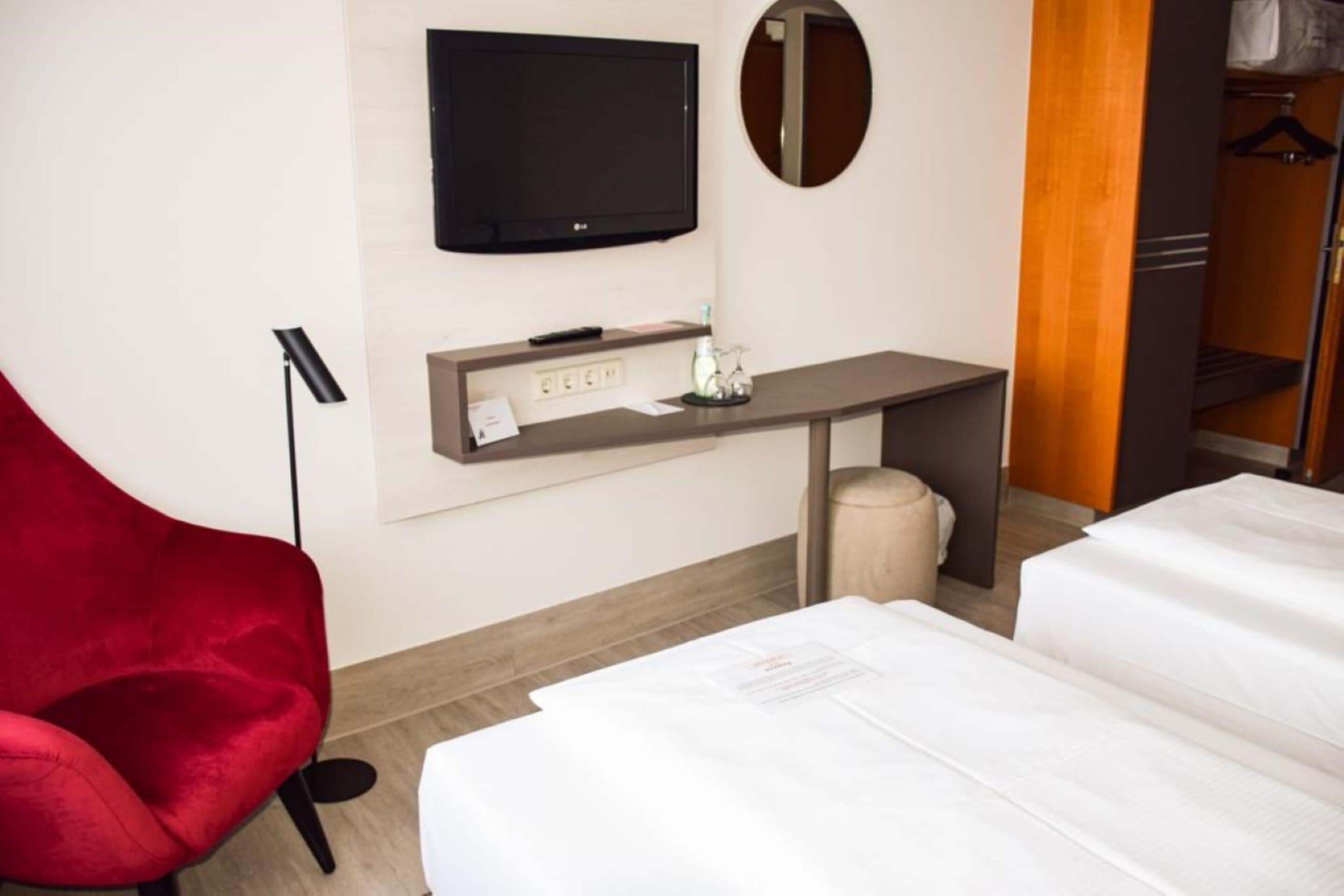 A hotel room with a bed, a desk with a flat-screen TV, a red chair, and a wardrobe.