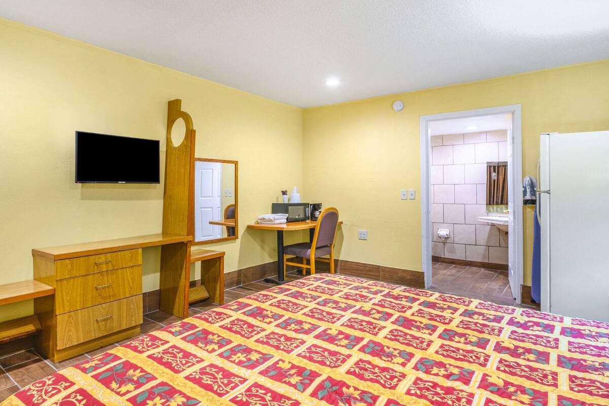 Quarto de hotel com cama, escrivaninha, cadeira, televisão, espelho e banheiro visível por uma porta aberta.