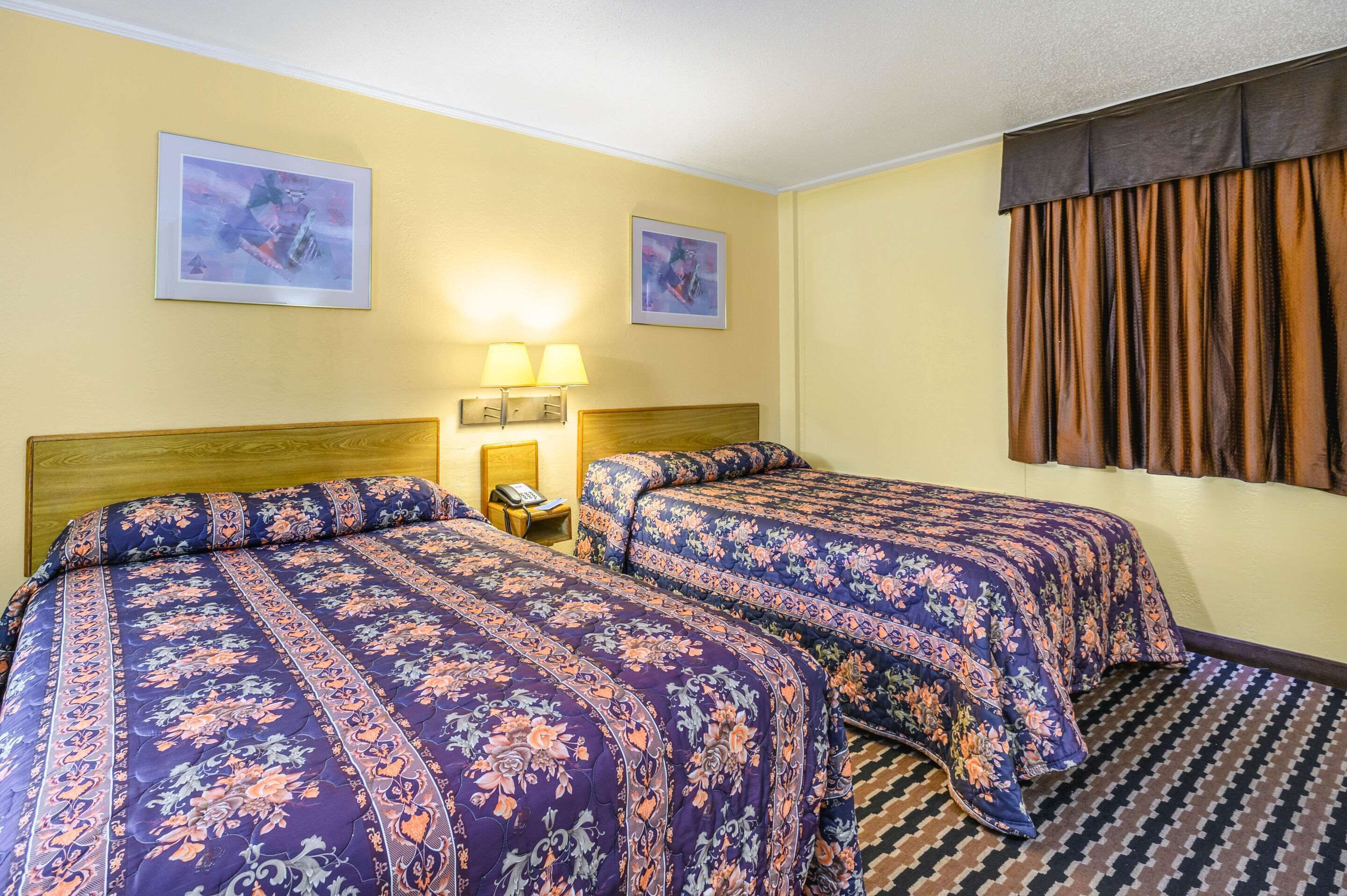 Ein Hotelzimmer mit zwei Betten, jeweils mit blumigem Bettbezug, einem Nachttisch mit Lampe und zwei gerahmten Bildern an den Wänden.