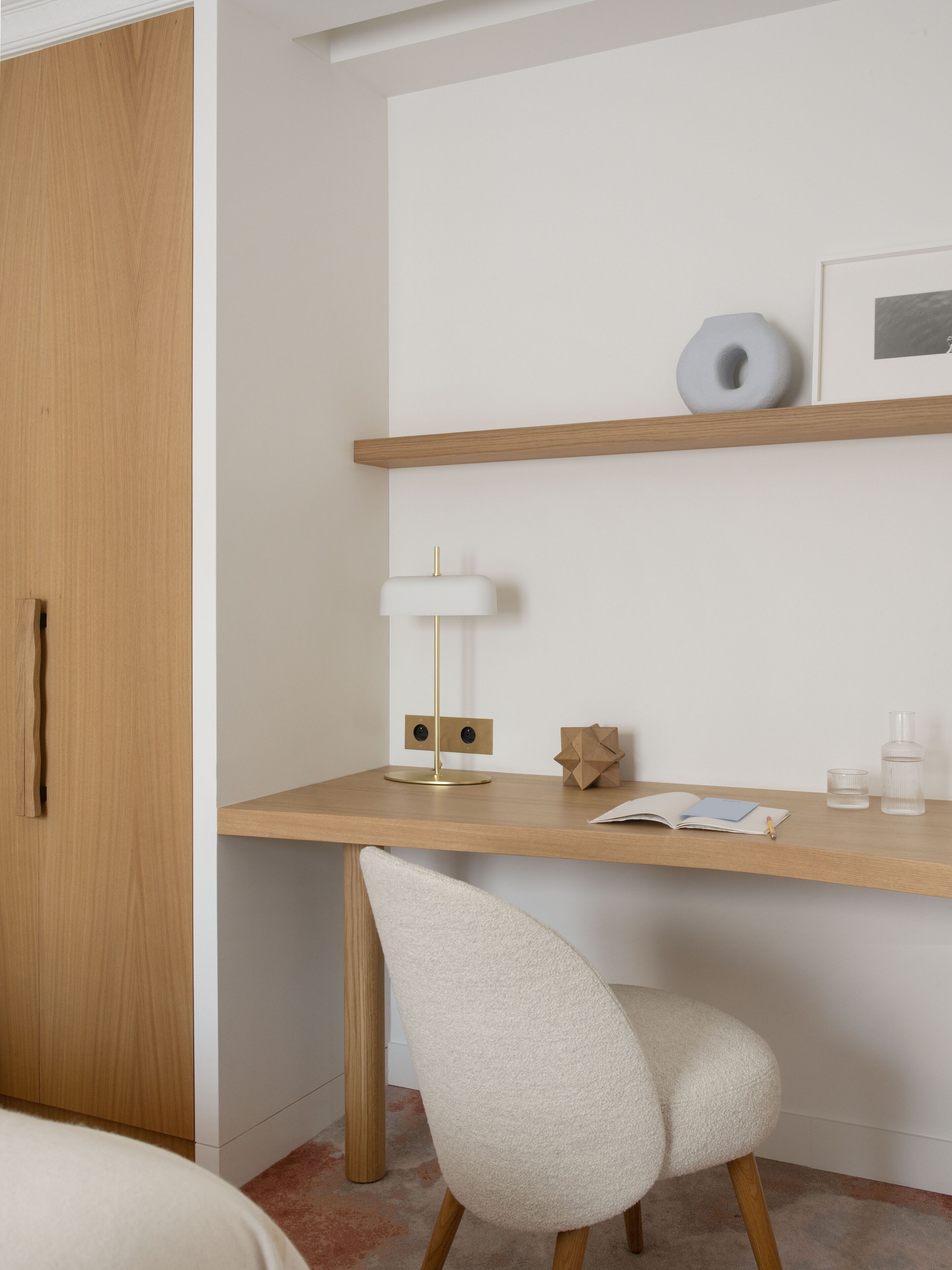 A modern room with a wooden desk, a white chair, a lamp, and a shelf with a picture and decorative items.