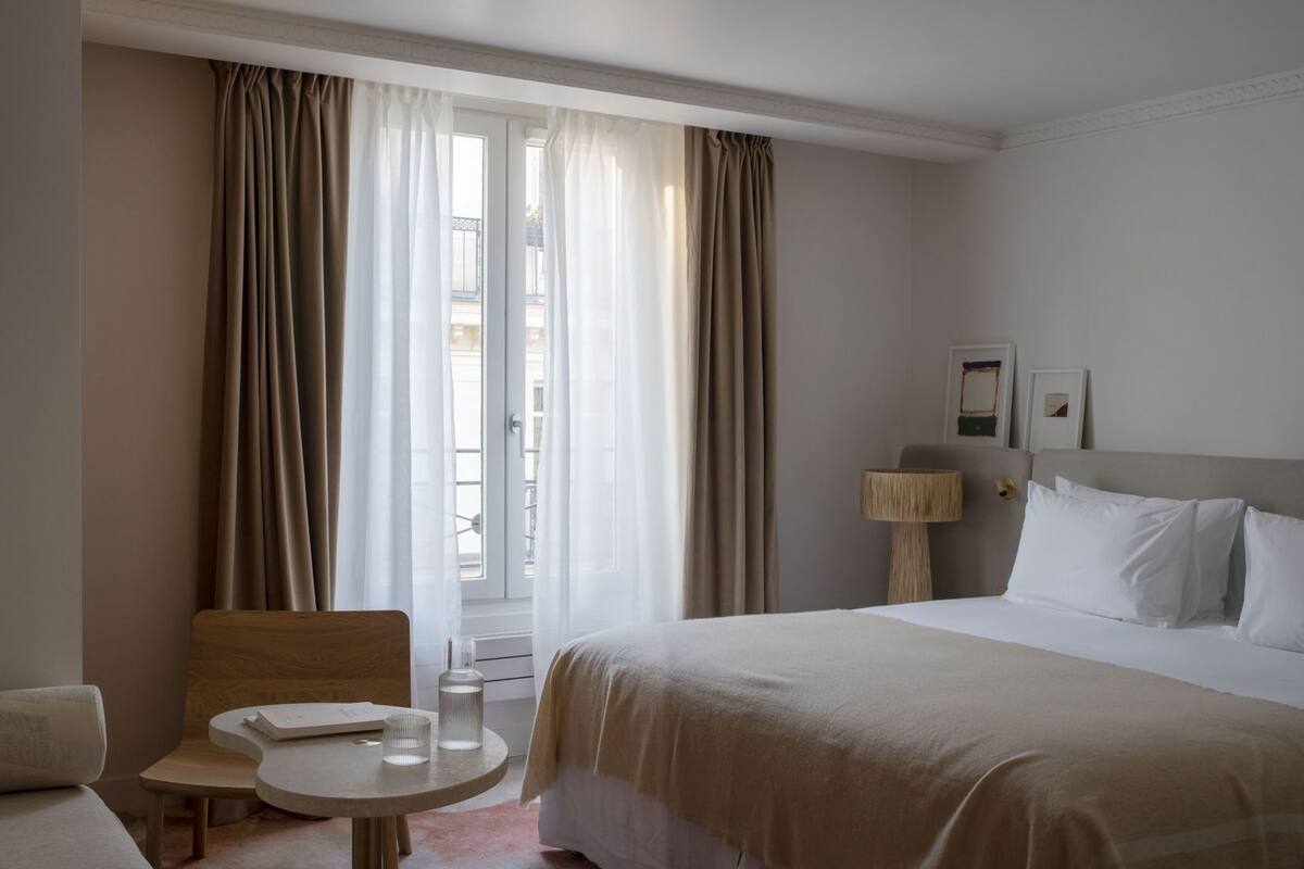 Una habitación de hotel con cama, una mesita, una silla, una lámpara y una ventana con cortinas.
