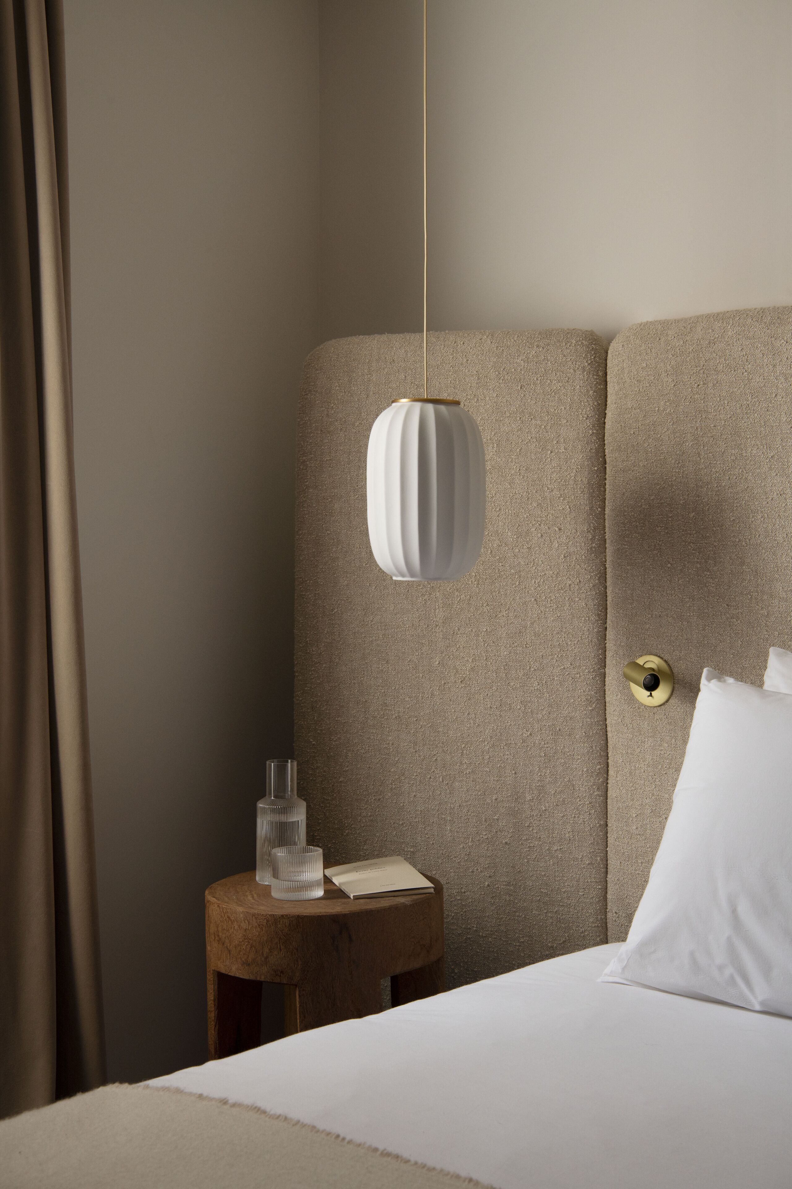 Cama bem arrumada com travesseiro branco e mesa de cabeceira de madeira ao lado. Uma luminária pendente branca paira sobre a mesa de cabeceira, onde se vê um frasco de vidro.
