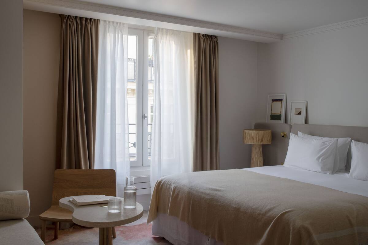 Habitación de hotel con cama, una mesita redonda con lámpara, una silla y un ventanal con cortinas.