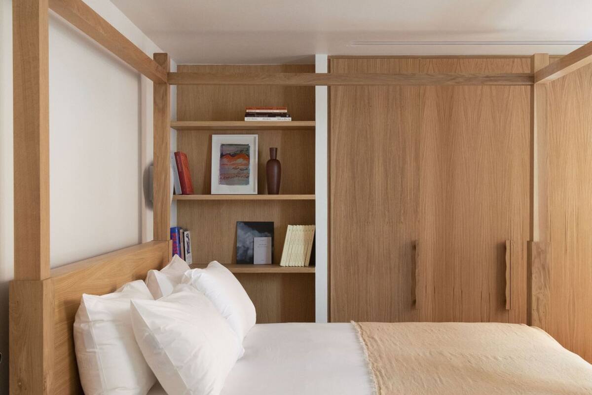 Un dormitorio con cama de madera, mesita de noche, armario y estante con libros y objetos decorativos.