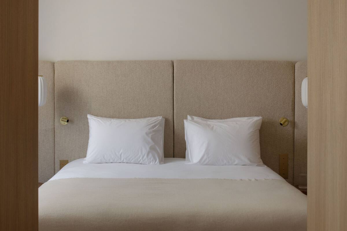Una cama doble con cabecera color beige y ropa de cama blanca, dos mesitas de noche con lámparas, y un marco de puerta de madera.