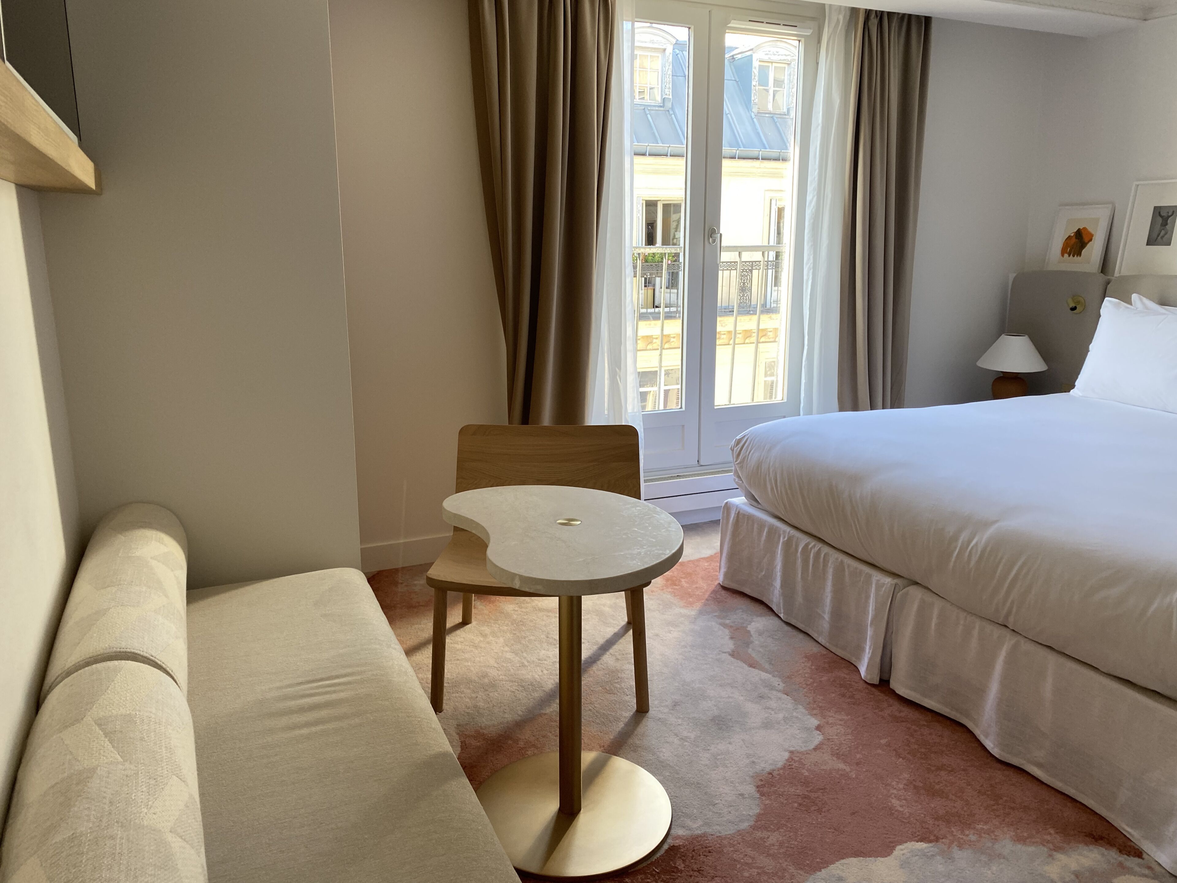 Ein Hotelzimmer mit einem Bett, einem kleinen runden Tisch, einem Sessel, einer Couch, einer Lampe und einem Fenster mit Vorhängen.