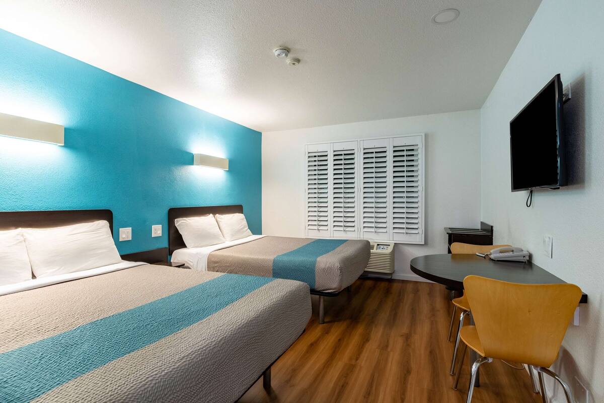 A hotel room with two beds, a TV, a desk with a chair, and a window with blinds.