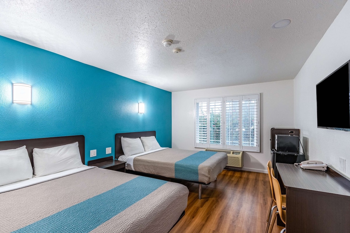 A hotel room with two beds, a desk, a TV, and a window with blinds.