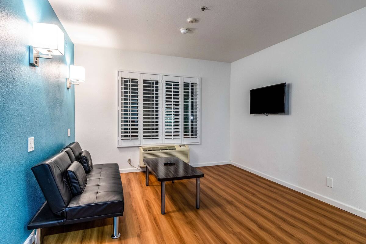 A room with a black leather sofa, a small table, a window with blinds, a mounted TV, and a ceiling vent.