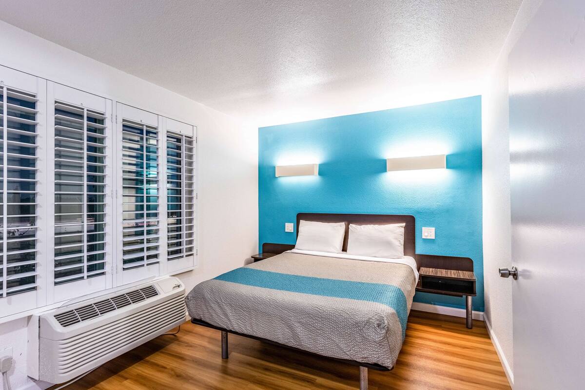 A bedroom with a bed, a window with blinds, a wall-mounted light fixture, and a heater.