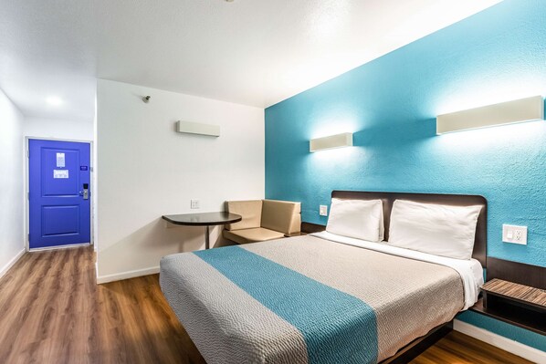 A hotel room with a bed, a small table, a chair, and a blue door.