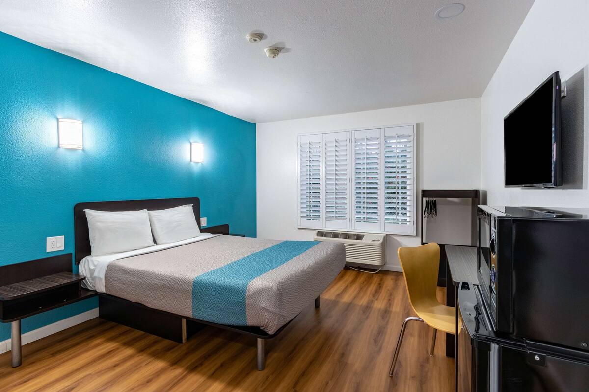 A hotel room with a bed, a desk, a chair, a TV, and a window with blinds.