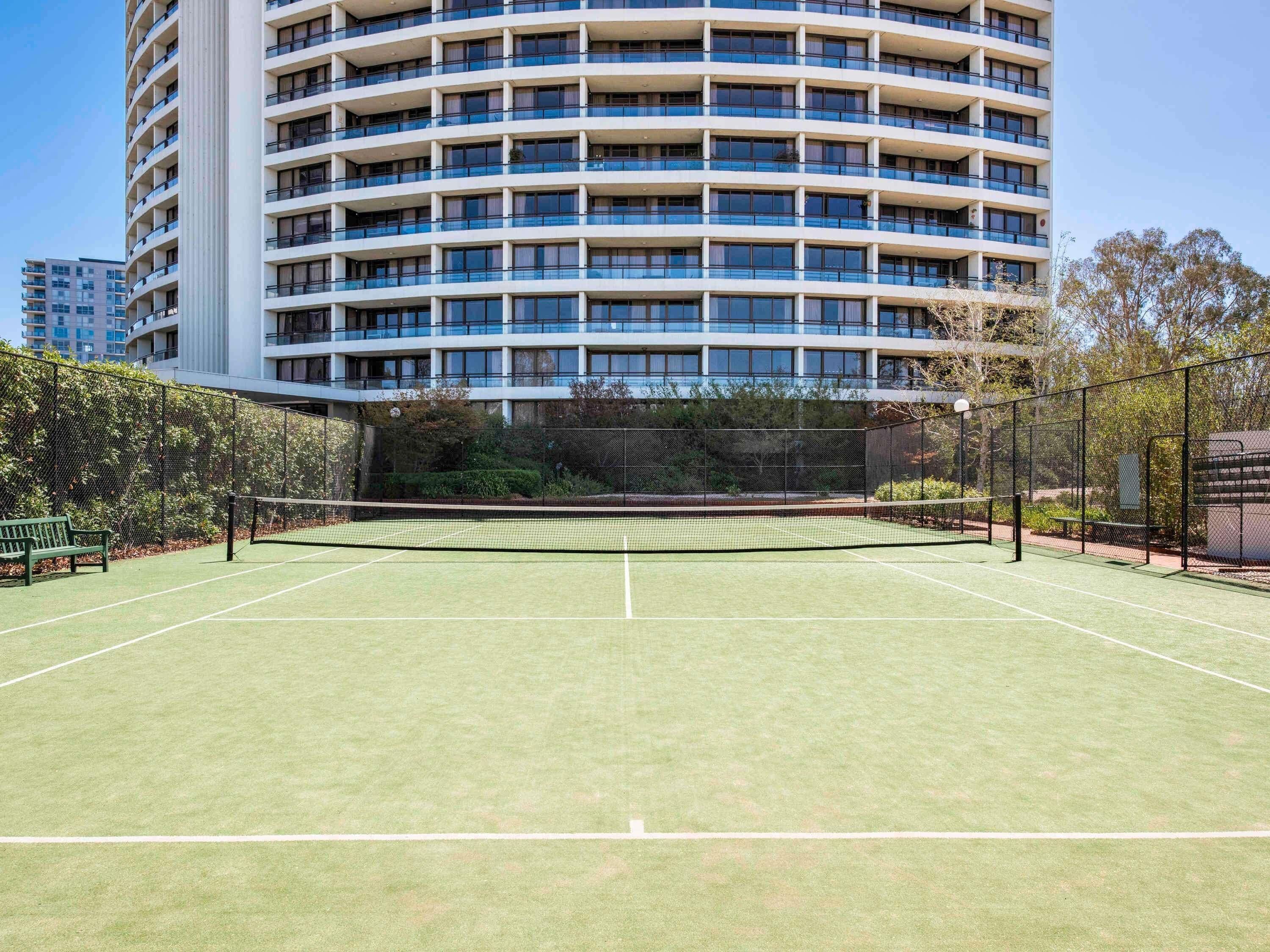 Un court de tennis avec un filet, entouré d’une clôture, et un bâtiment avec de nombreux balcons en arrière-plan.