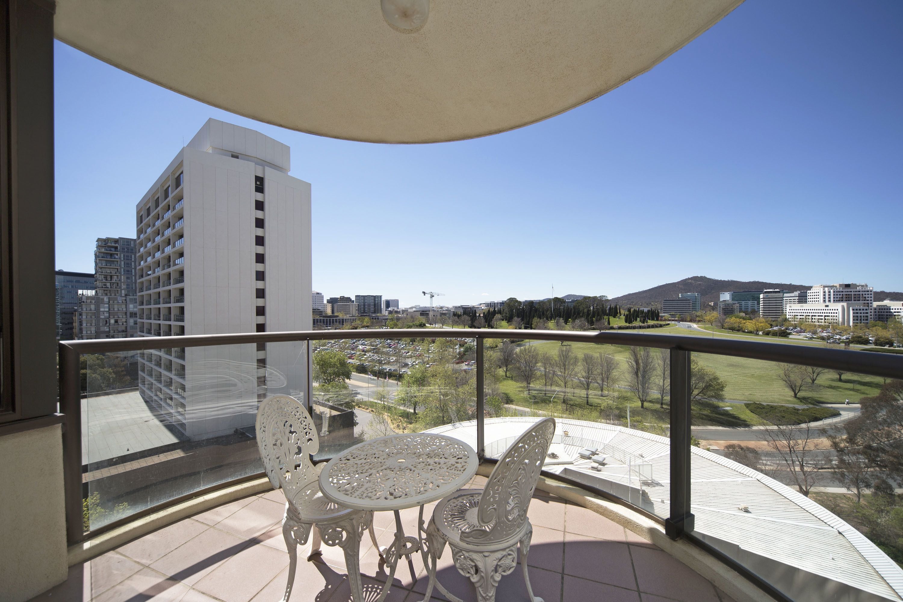 A balcony with a table and chairs, offering a view of a parking lot, buildings, and a park.