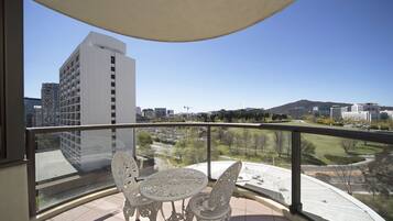 A balcony with a table and chairs, offering a view of a parking lot, buildings, and a park.